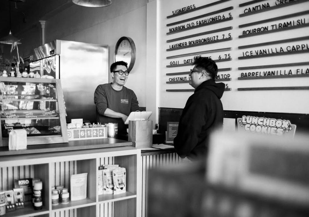 A cashier and a customer in a coffee shop with shelves and a counter.