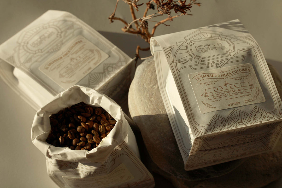 2 12oz bags of sealed coffee beans on brances and rocks and one bag open revealing coffee beans
