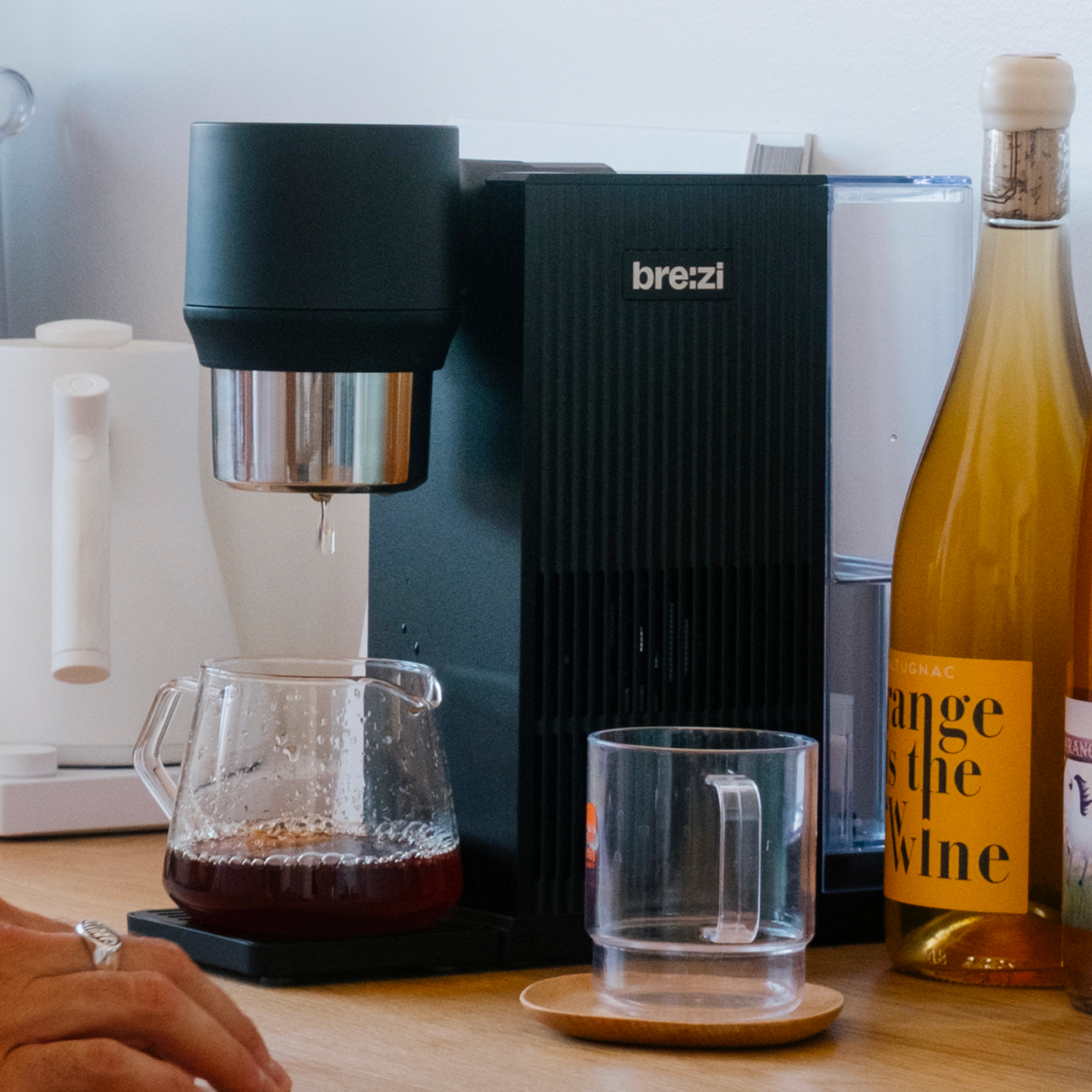 Brezi coffee machine on a wooden table with a glass of water and a bottle of orange juice.