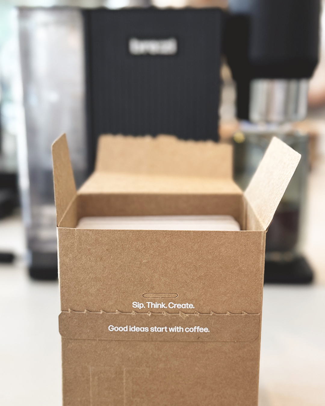 Open cardboard box with coffee pouches inside, blurred coffee machine in the background