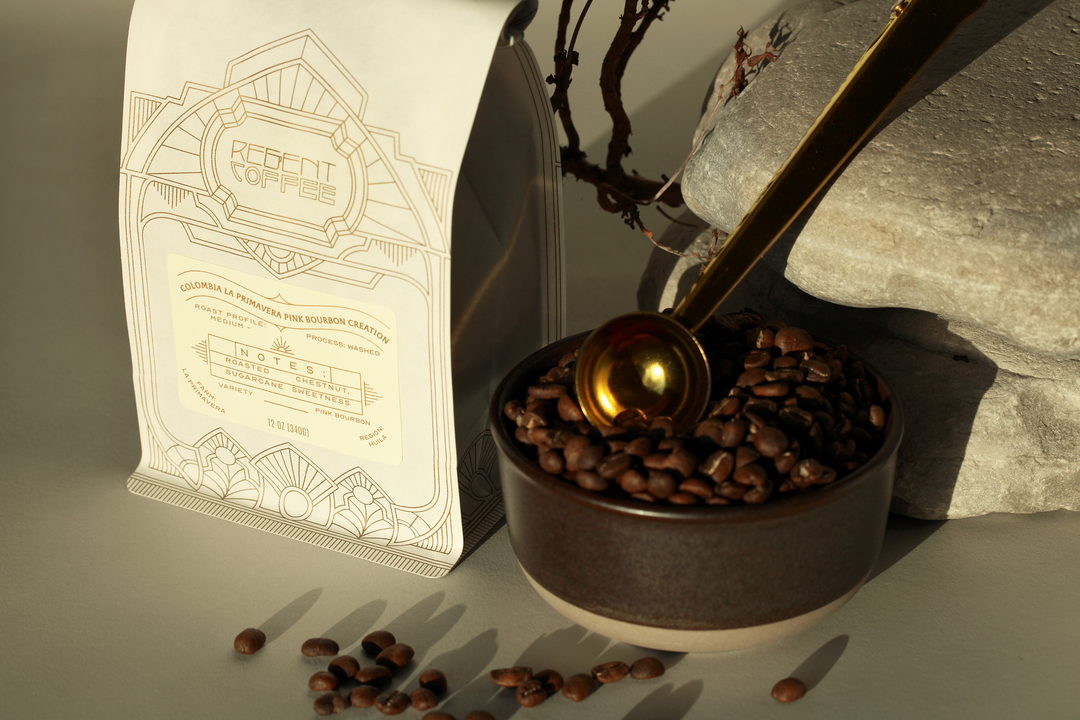 12oz bag of Colombia La Primavera Pink Bourbon Creation on the left and a bowl with coffee beans and a gold spoon resting on a rock on the right
