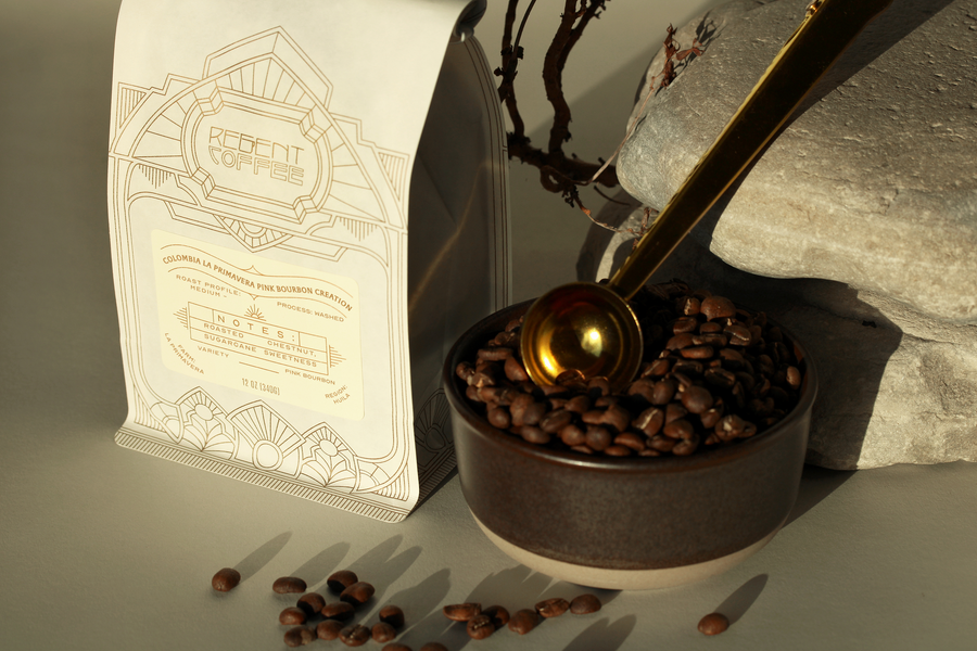 12oz bag of Colombia La Primavera Pink Bourbon Creation on the left and a bowl with coffee beans and a gold spoon resting on a rock on the right
