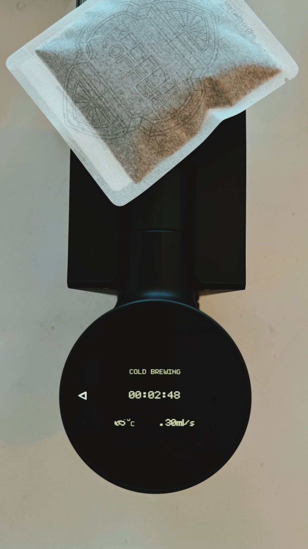 A close-up of a black cold brew coffee maker with a digital display showing brewing progress, topped with a paper filter bag containing coffee grounds on a light beige background.