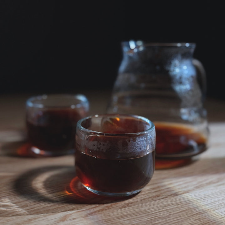Two glass cups filled with a dark liquid, possibly coffee or tea, on a wooden surface with a blurred background.