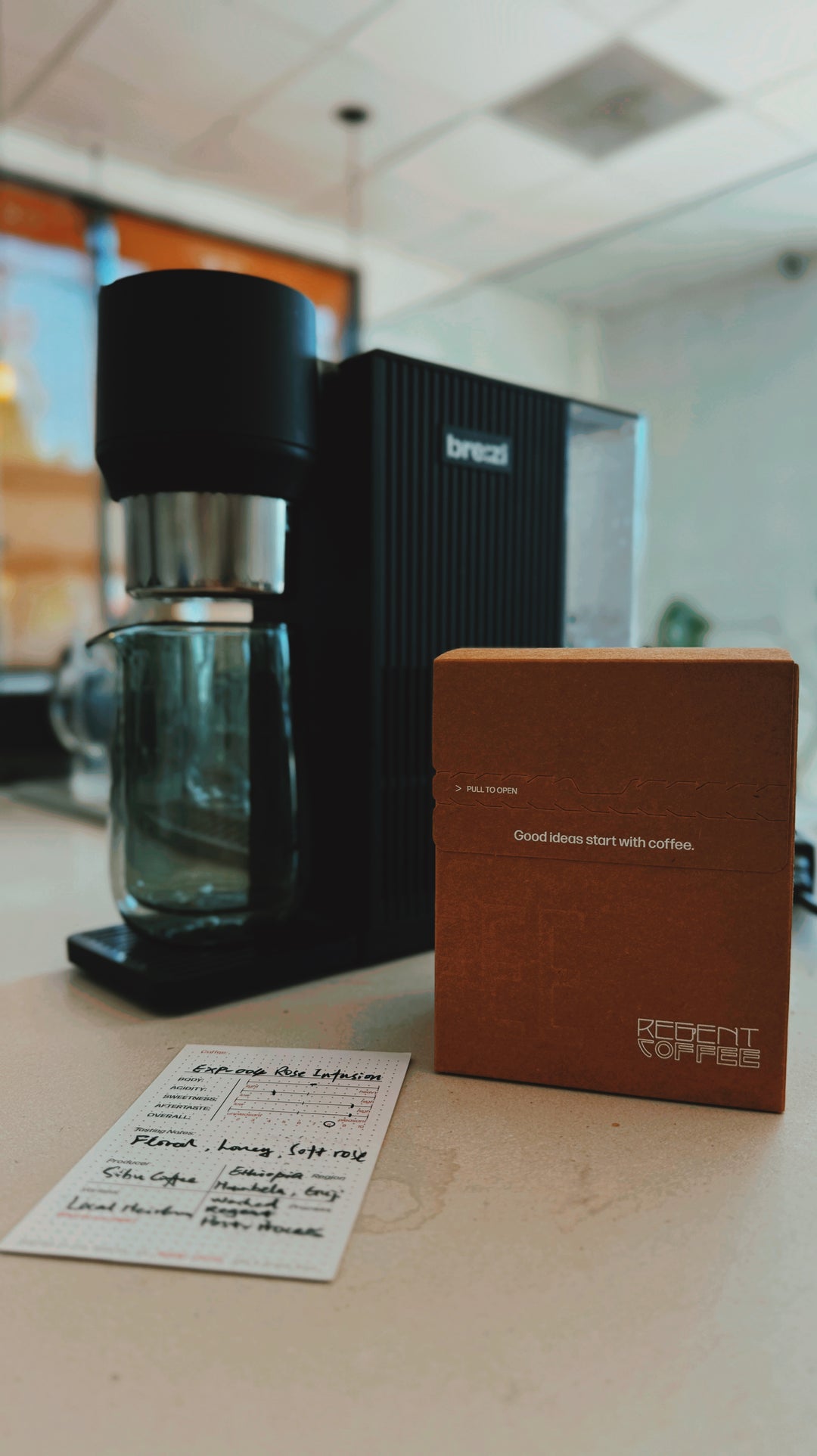 A coffee maker with a black and silver body, a black vertical coffee machine, a brown cardboard box, and a handwritten paper card on a light countertop in a kitchen setting.
